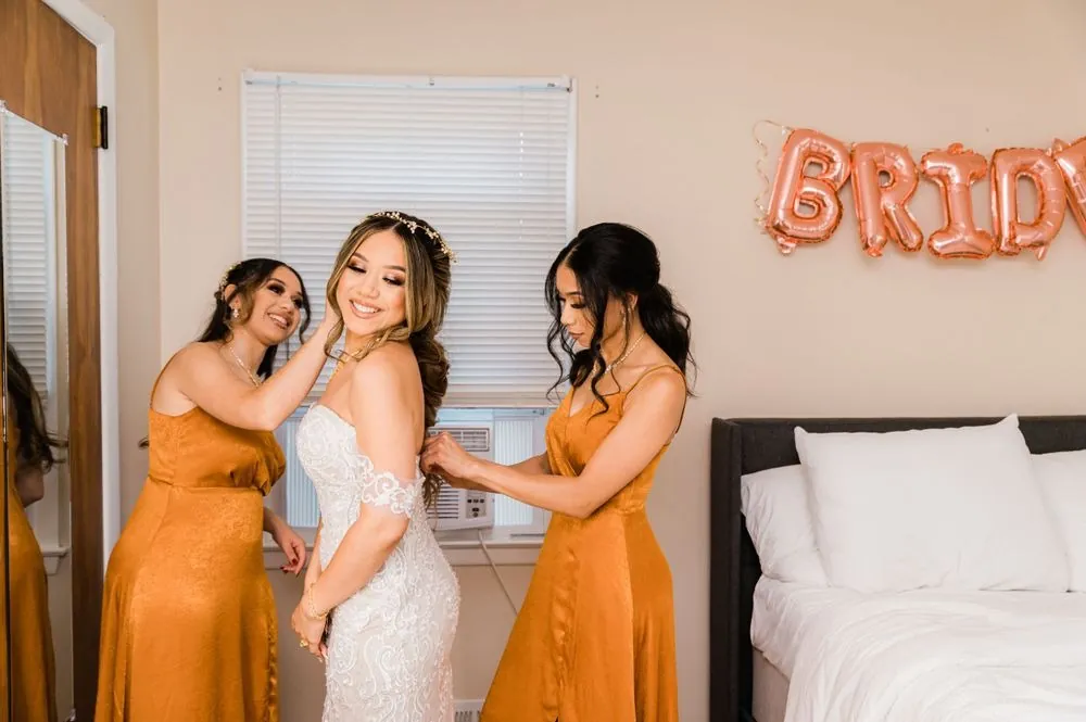 Bridesmaids in burnt orange satin fasten the bride's gown under a BRIDE balloon banner
