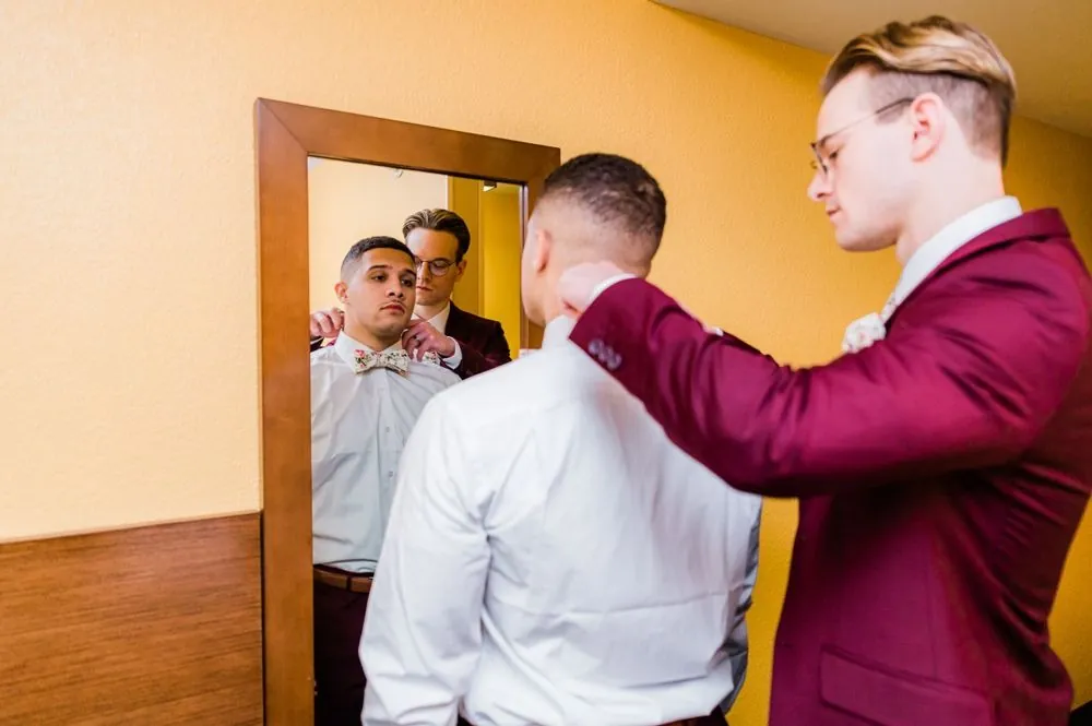 A groomsman in burgundy straightens the groom's collar in front of a mirror