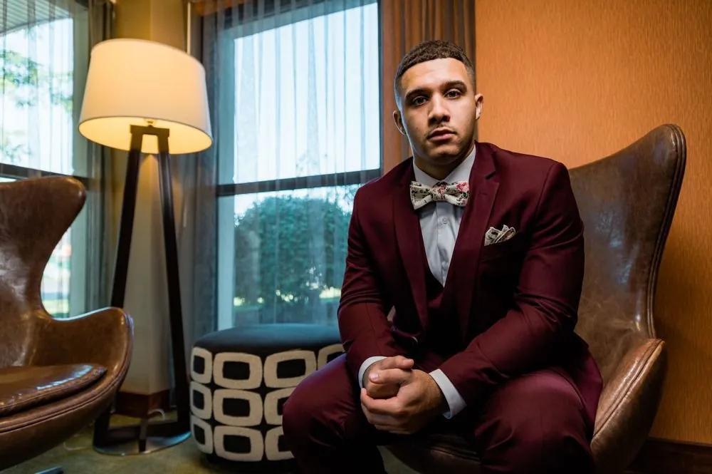 The groom in a burgundy suit sits in a leather chair beside a sunny window