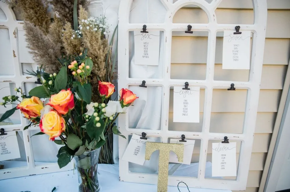 A white window frame seating chart with orange roses, pampas grass and a gold T