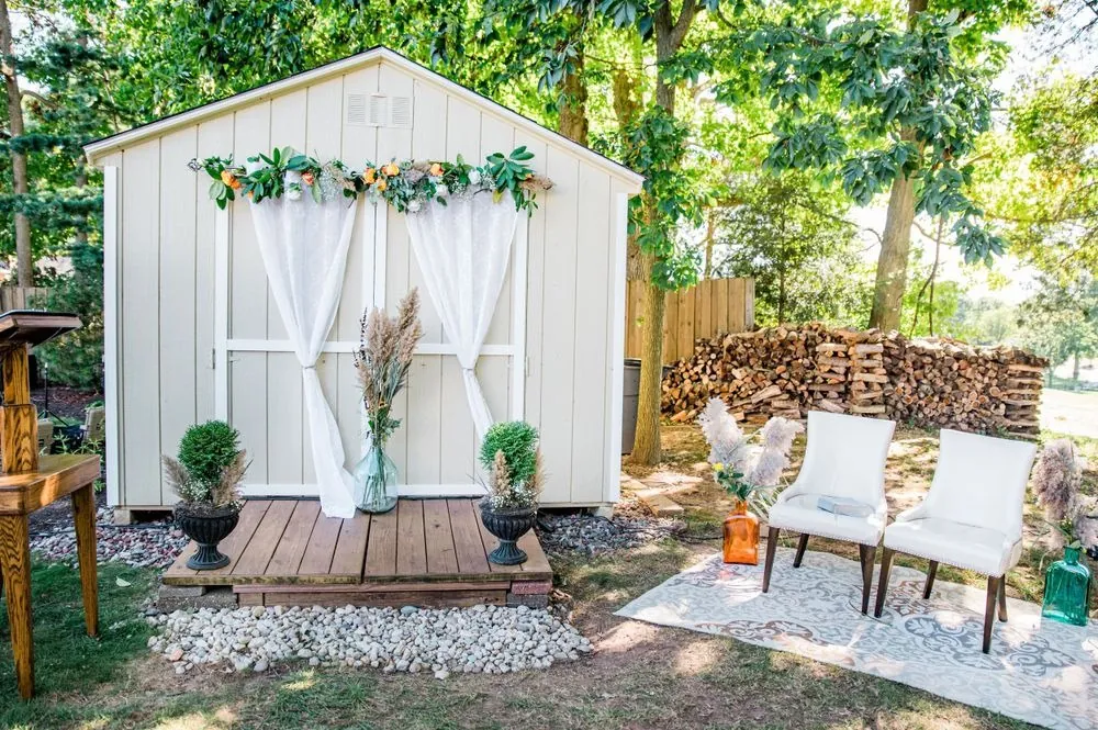 A backyard shed dressed with sheer curtains, a floral garland and 2 white chairs