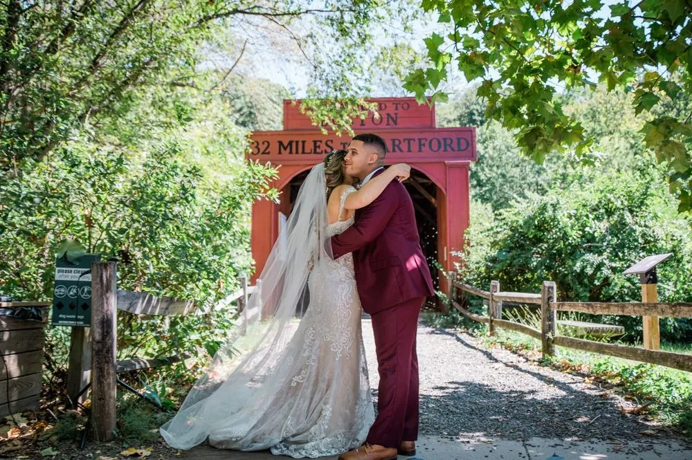Tatiana and Tyler hug on the gravel path in front of a red covered bridge
