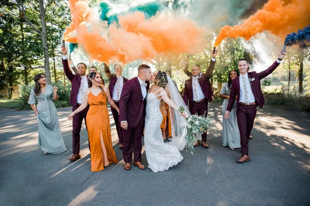 The wedding party cheers behind the couple as orange and teal smoke fills the air
