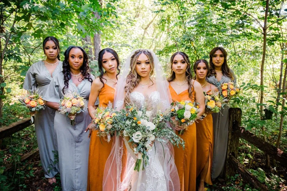 Bridesmaids in sage and marigold gowns surround the bride among green trees