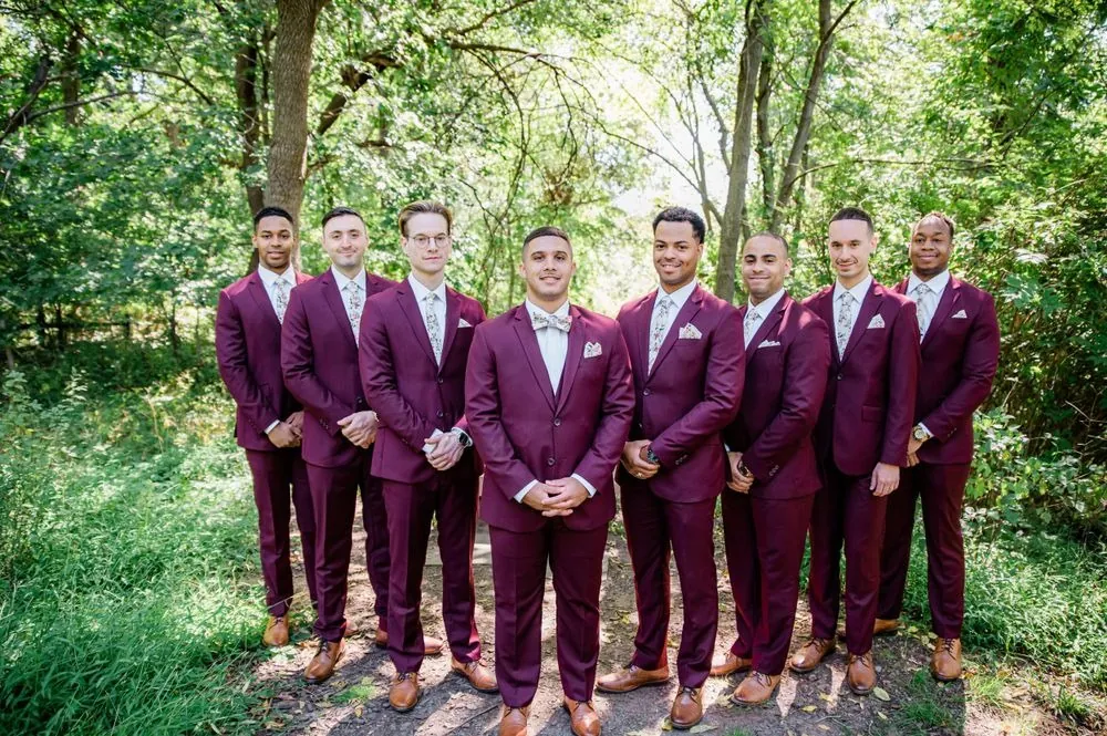 Groomsmen in burgundy suits and floral bow ties line up in the woods