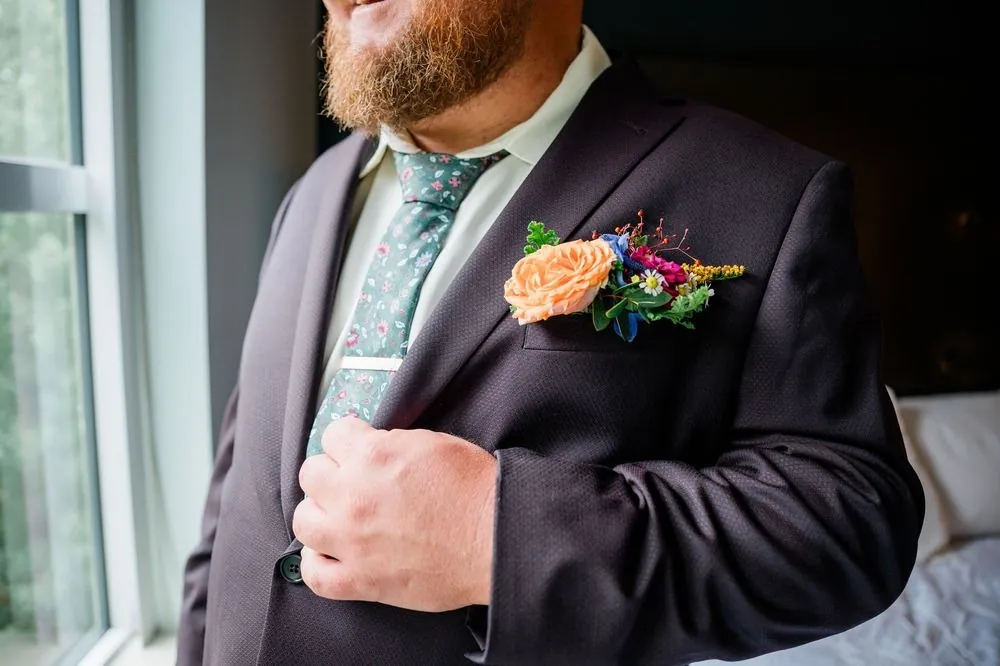 The groom buttons his eggplant suit jacket, a bright boutonniere on the lapel