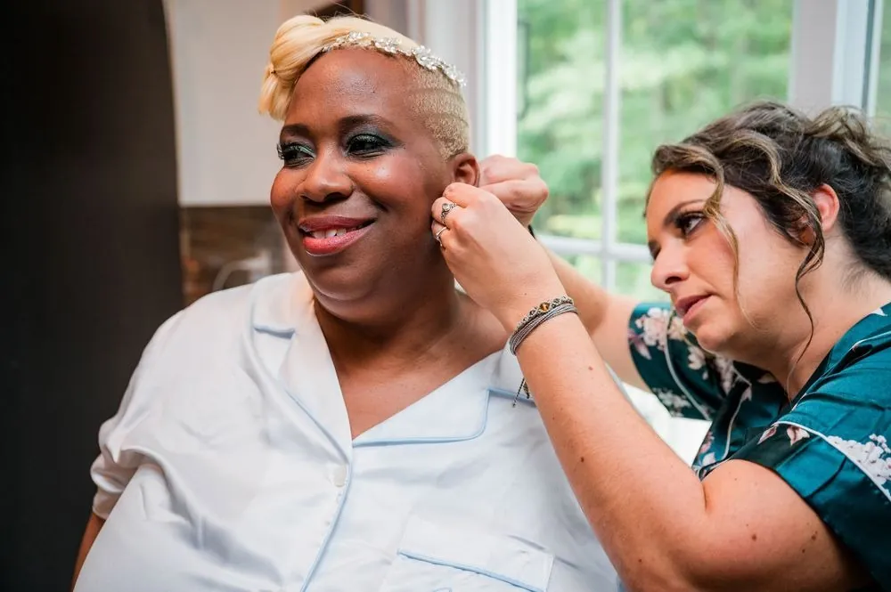 A friend fastens the bride's earring while she smiles near the window
