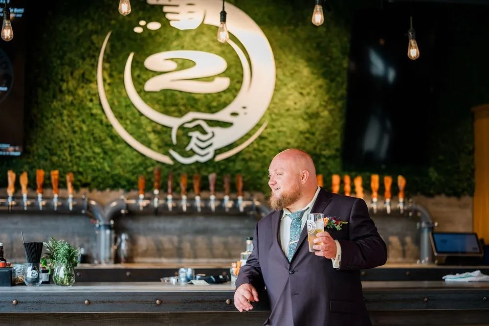 The groom stands at the tap wall under the Two Roads logo, beer in hand