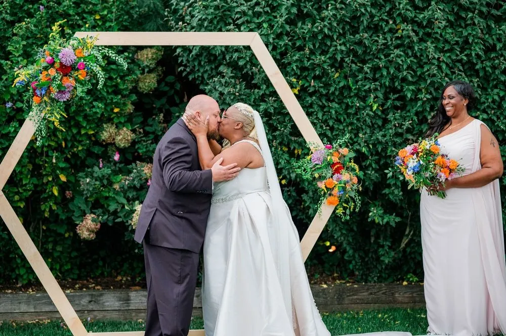 Tiffanie and Kevin kiss under the flowered hexagon arch against a green hedge