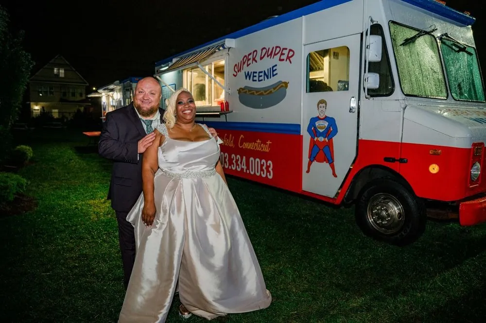 Tiffanie and Kevin stand on the grass beside the lit hot dog truck at night