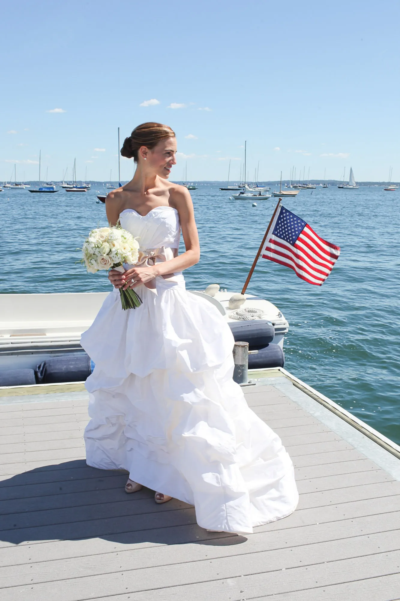 The bride stands on the dock in a ruffled gown with an American flag and sailboats behind