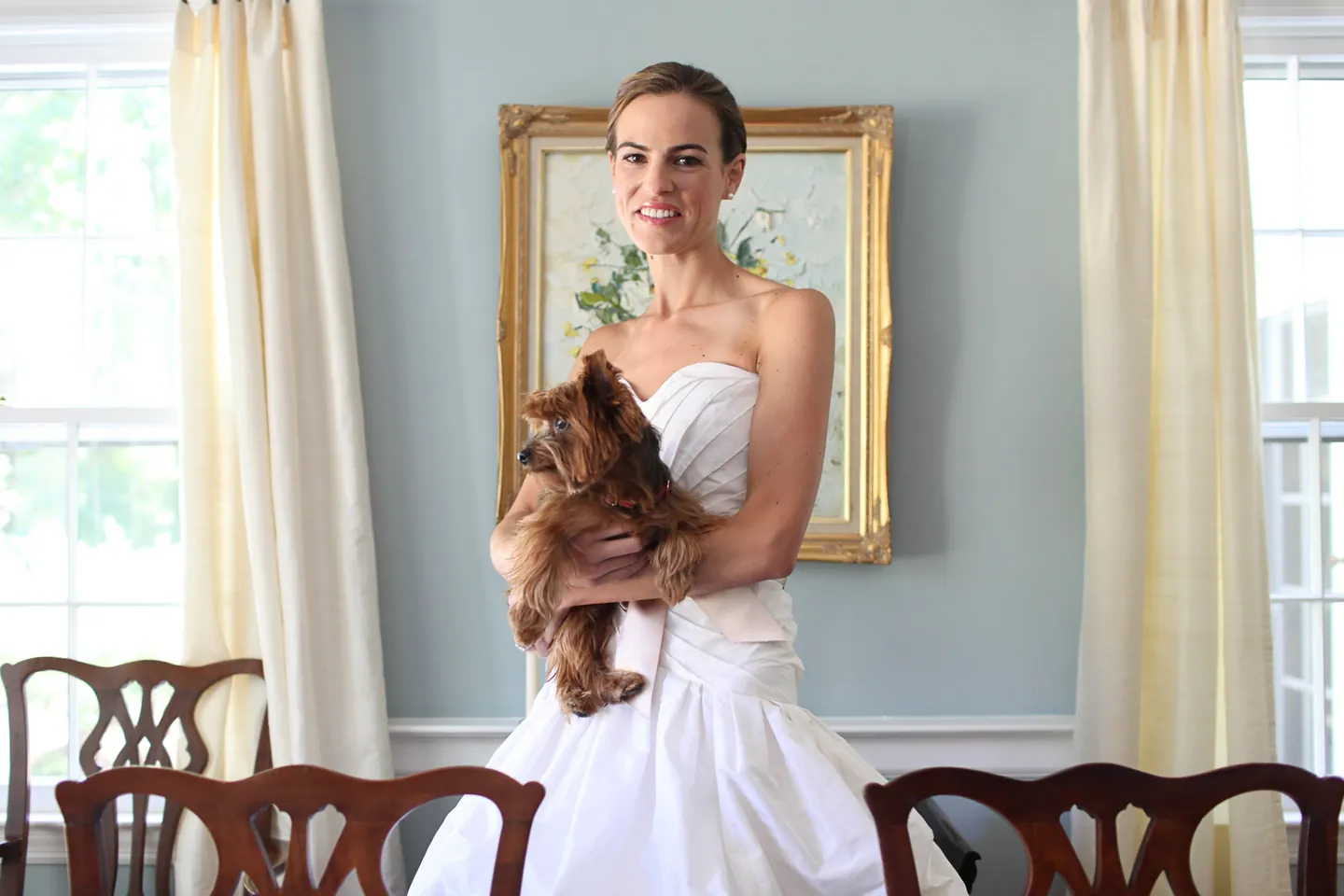 The bride in a strapless gown cradles a small brown dog beside a framed painting and curtains