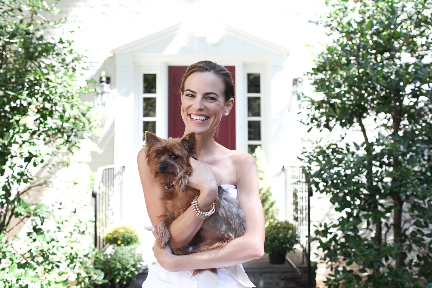 The bride laughs holding a small brown dog in front of a white building with a red door