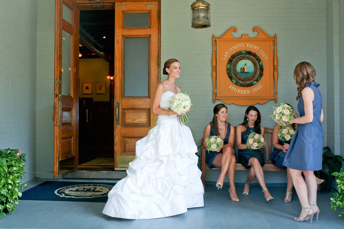 The bride steps out of the club doorway as bridesmaids in navy wait with bouquets