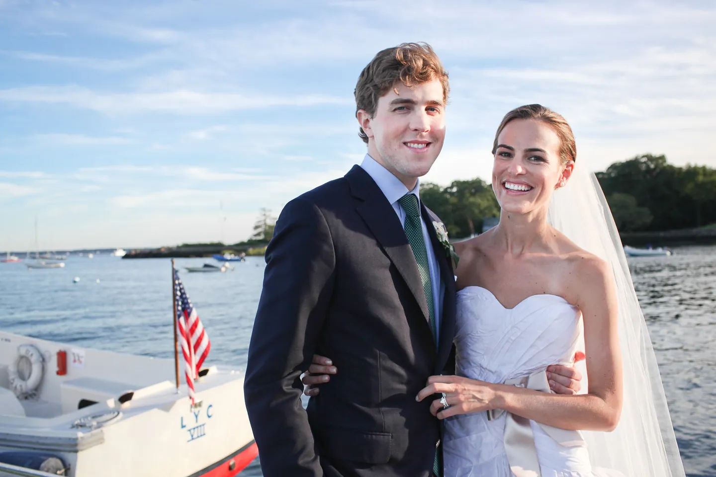 Trevor and Christina smile on the dock with the harbor and a flag-flying launch behind