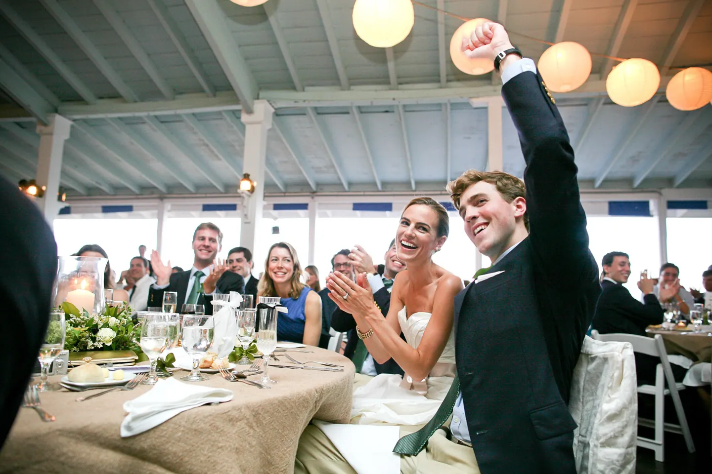 The groom throws a fist in the air at the head table as the bride laughs