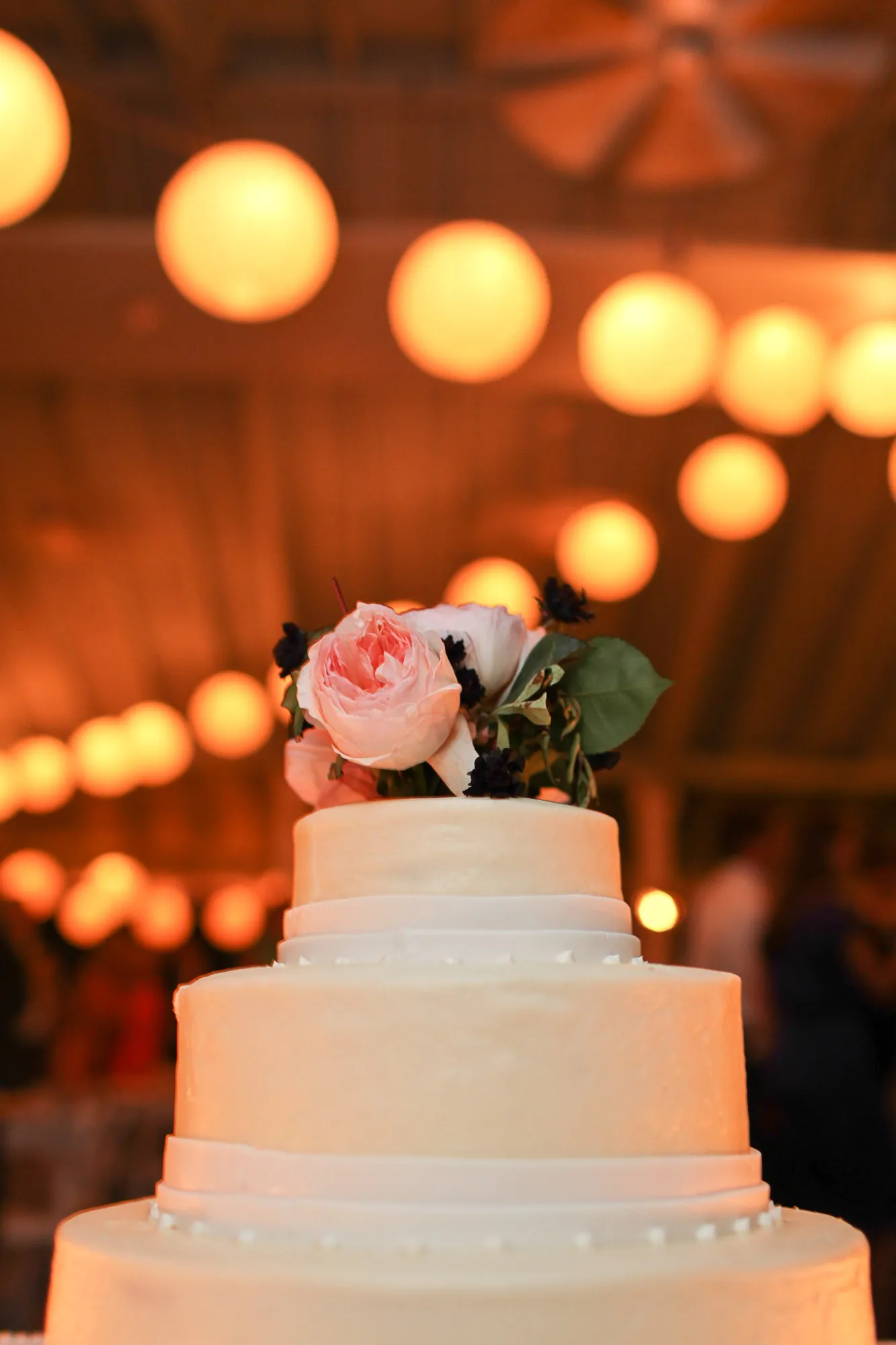 A tiered white wedding cake with a pink flower on top under warm lanterns