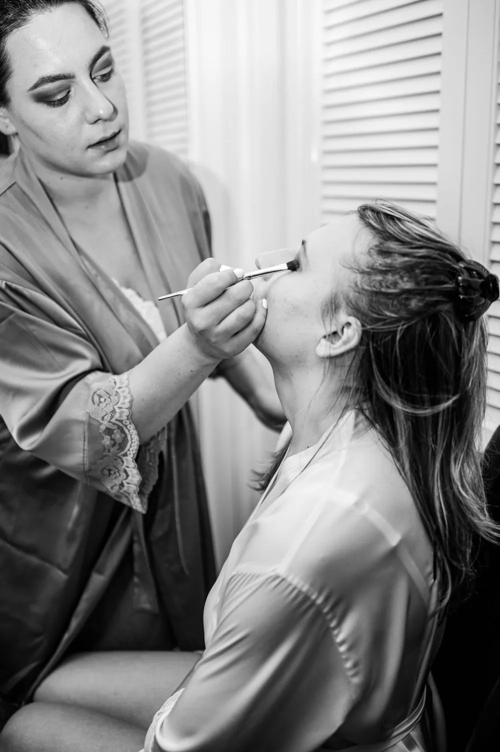 Black and white frame of the bride having her makeup done by the window