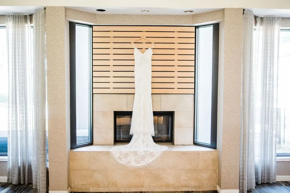 The lace wedding gown hangs in a tall window above a fireplace