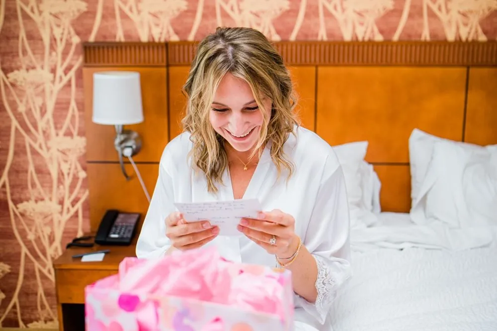 The bride smiles while reading a card on the bed in her white robe