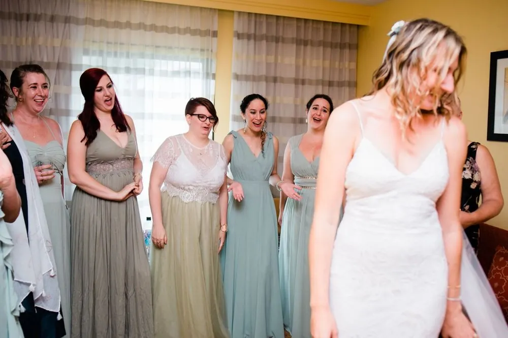 Bridesmaids in sage dresses react as the bride turns in her gown