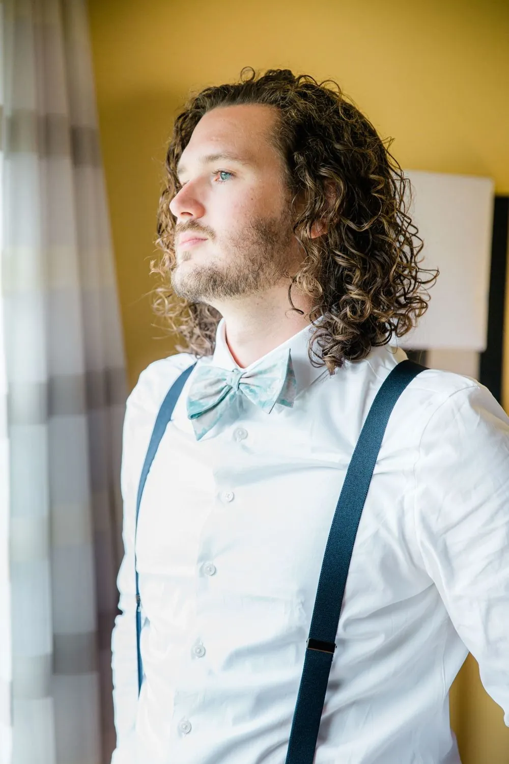 The groom with long curly hair adjusts his suspenders by a window
