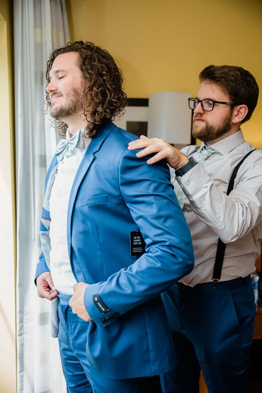 A groomsman helps the groom into his blue suit jacket