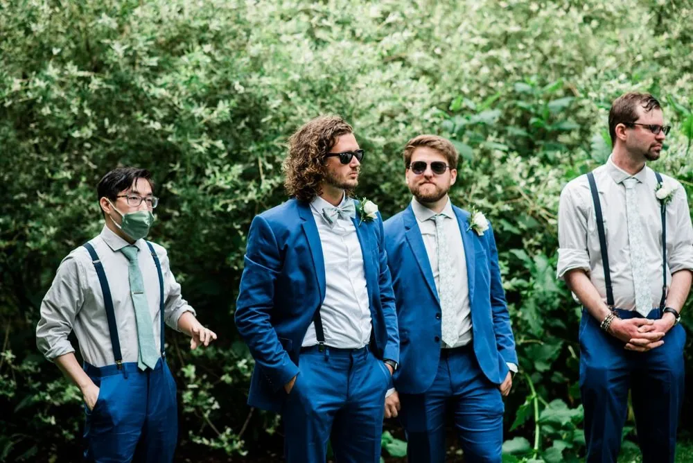 The groom and groomsmen in blue and suspenders stand in the garden greenery