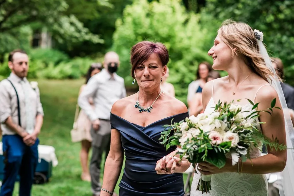 A woman in navy walks the bride into the garden ceremony past masked guests