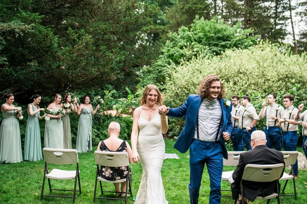 Vita and Mat walk hand in hand past white chairs as the wedding party cheers