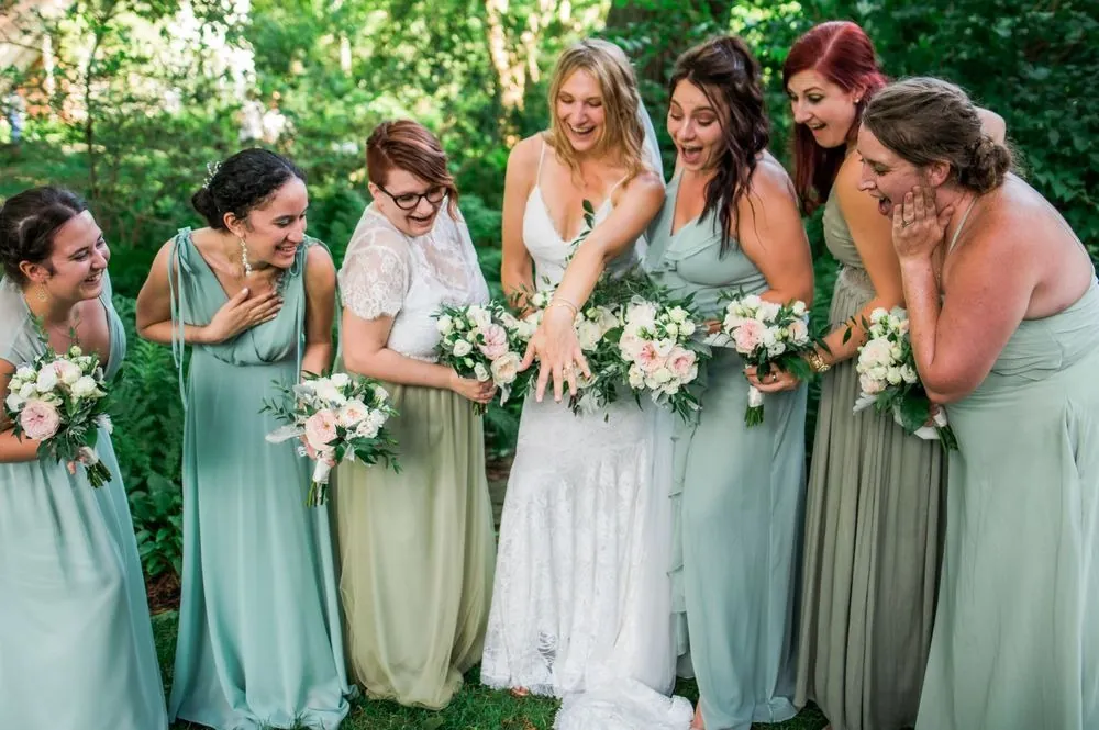 The bride laughs with bridesmaids in sage gowns holding white bouquets