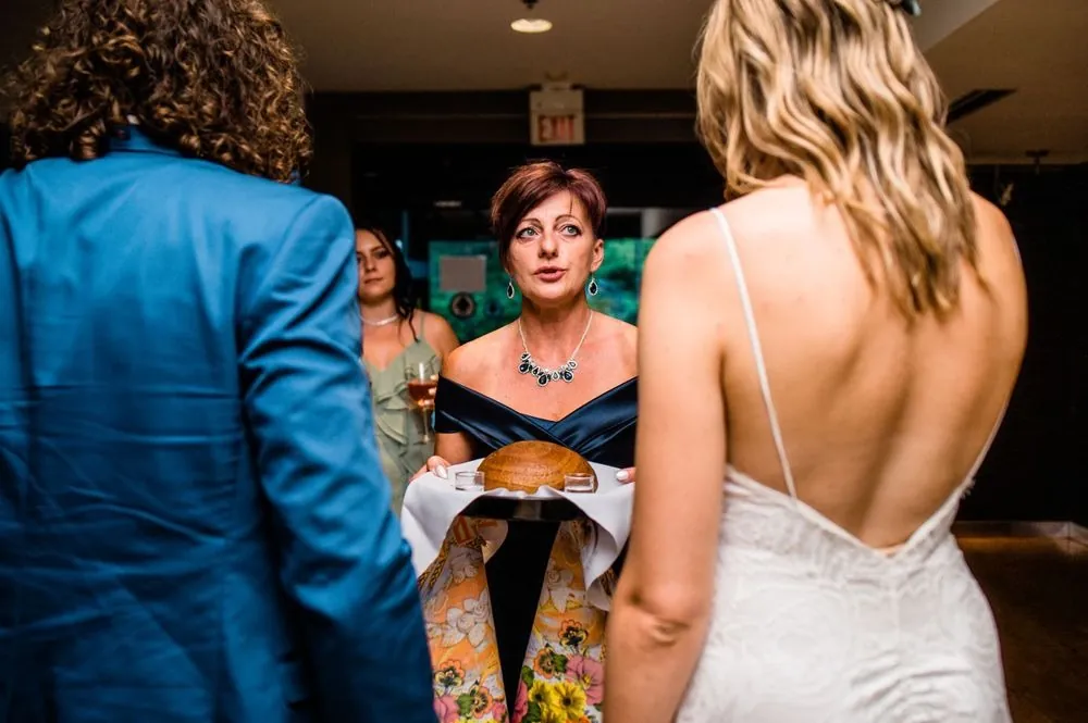 A round loaf on an embroidered towel is presented to the couple indoors