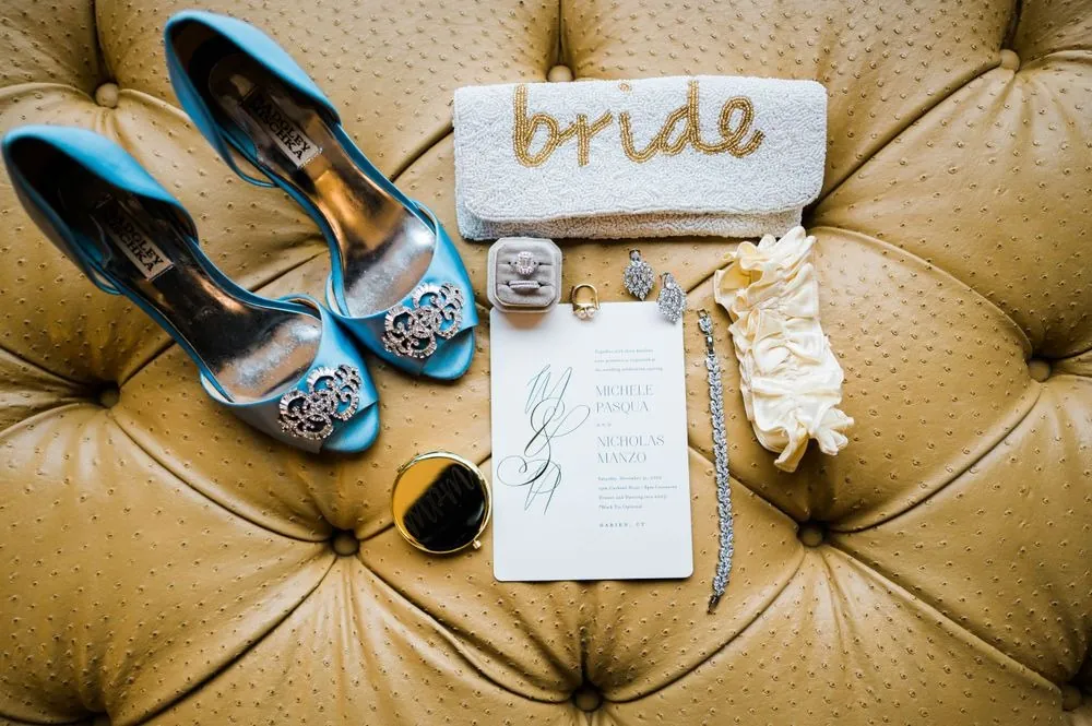 Teal satin heels, a beaded bride clutch, and invitation laid on a tufted leather chair