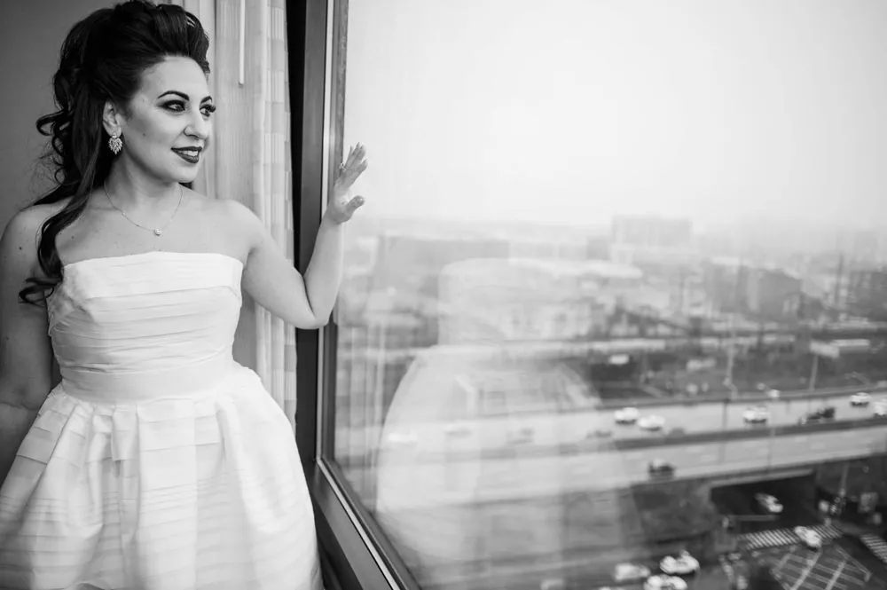 Black and white frame of the bride in her gown looking out a tall hotel window
