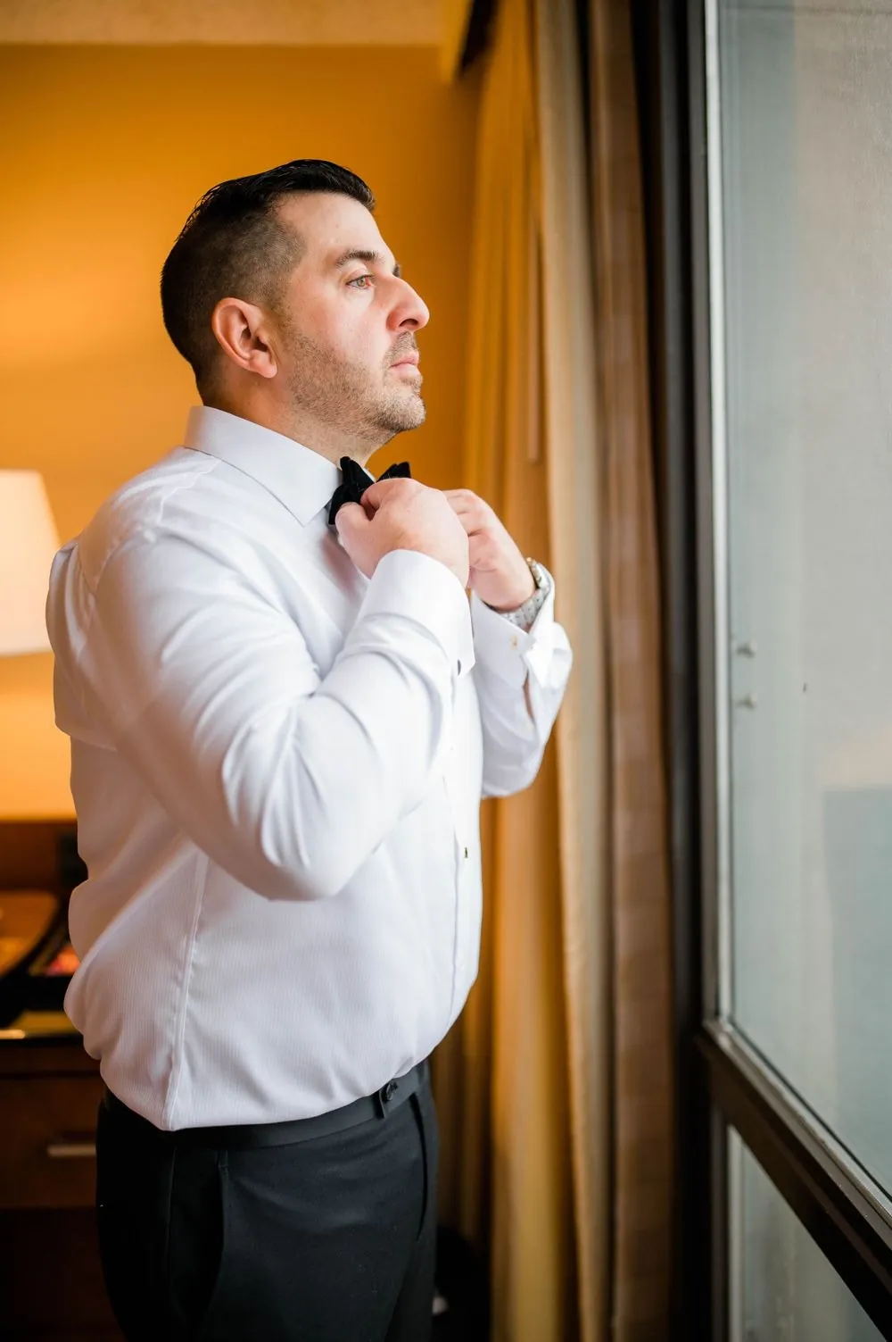 Groom in a white shirt adjusts his sleeve beside a window with sheer curtains