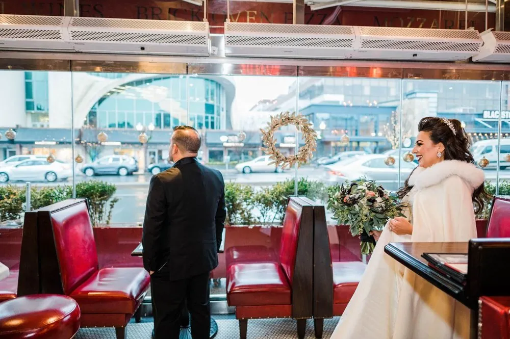 Bride in a white fur wrap walks up behind the groom by red booths in a pizzeria
