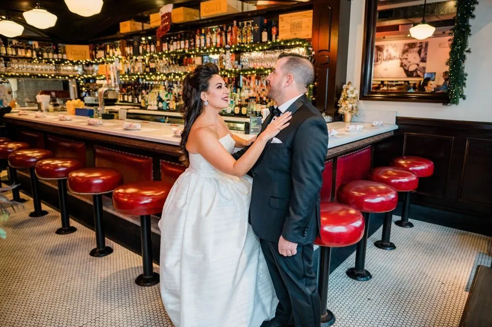 Michele and Nicholas laugh against a marble counter lined with red stools and holiday garland