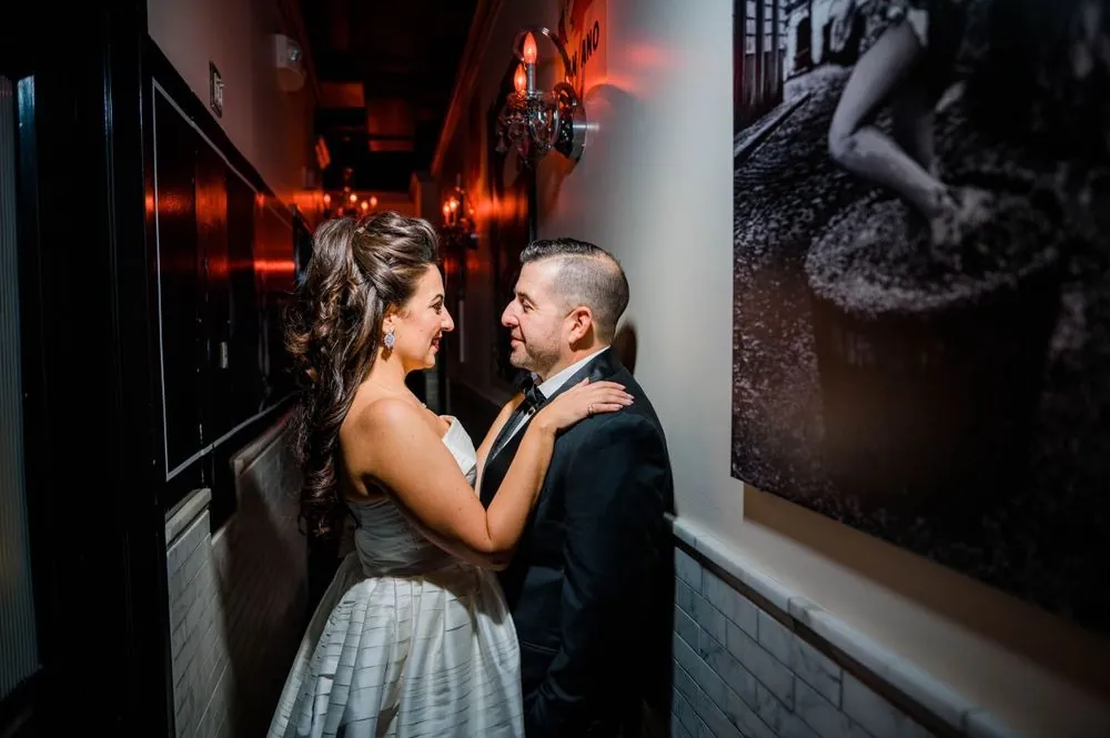 Michele rests her hand on Nicholas's chest in a dim hallway lit by red sconces