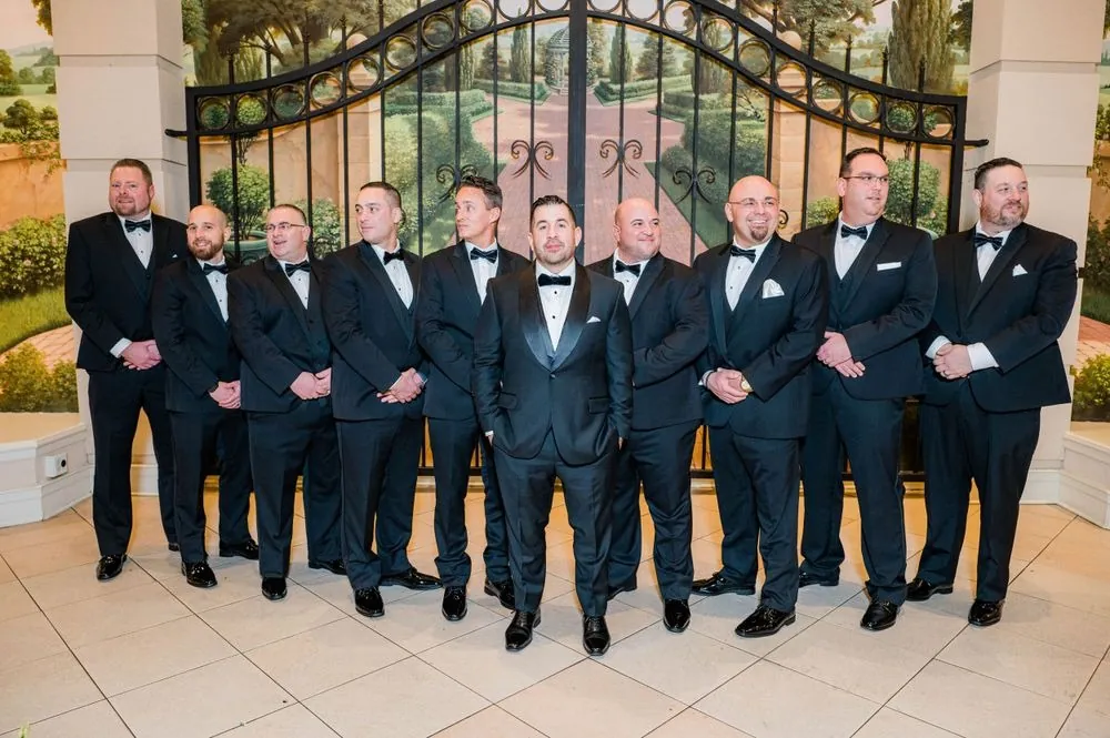 Groom and groomsmen in black tuxedos stand in a line before a painted garden mural