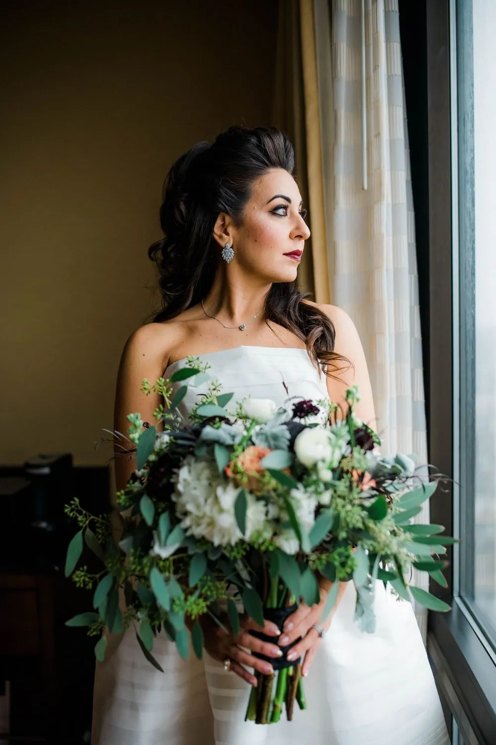Bride holds a bouquet of white roses and greenery by a curtained window