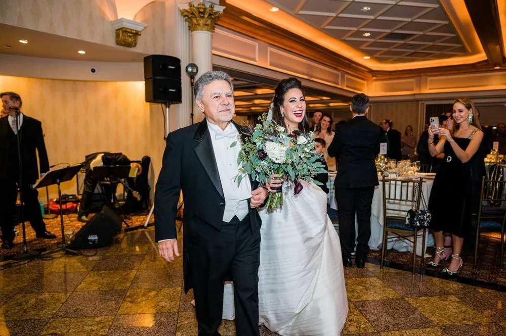 Older man in a tuxedo walks the bride into the ballroom past seated guests