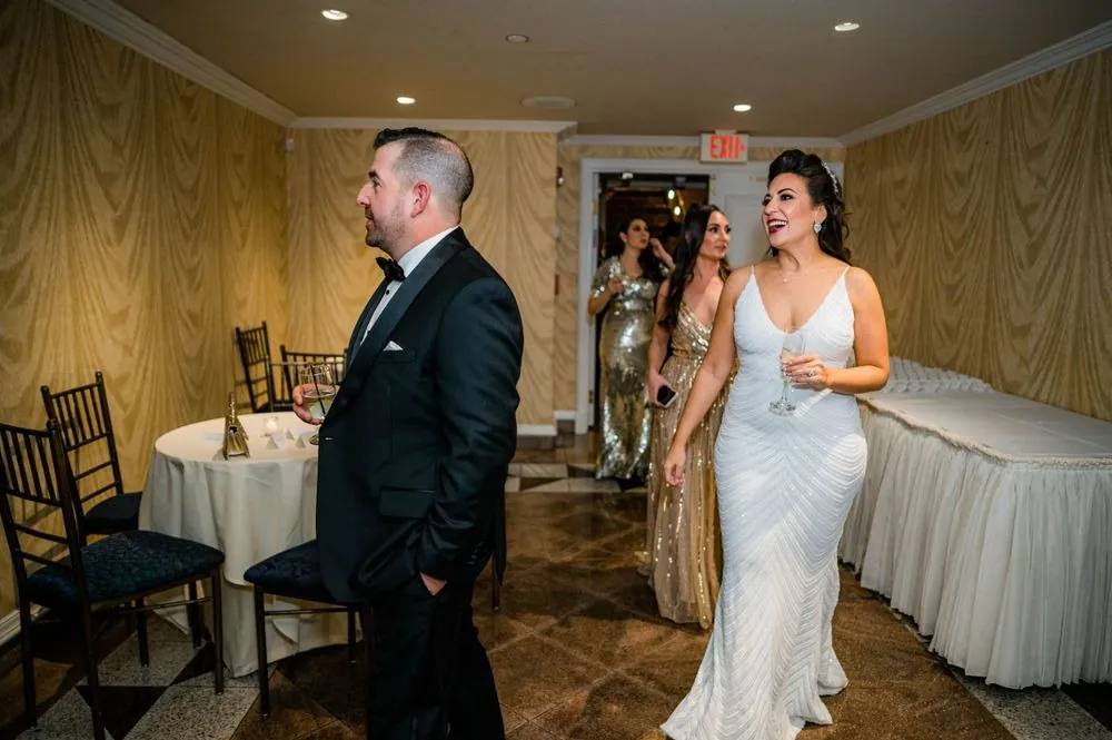 Bride in a fitted beaded gown walks in holding a drink as the groom watches