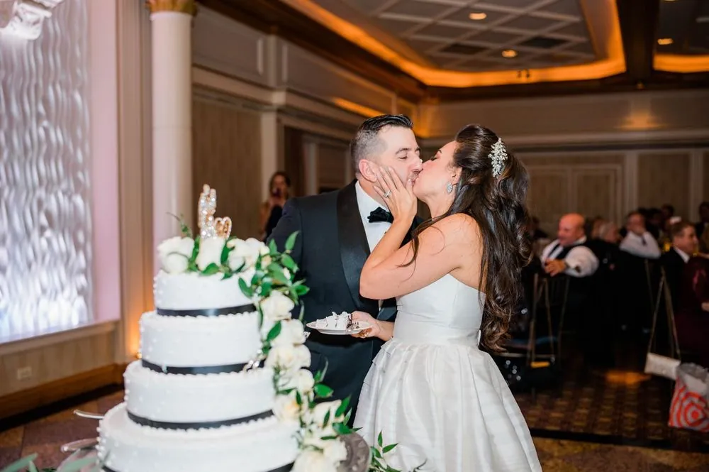 Michele and Nicholas kiss behind a white tiered cake trimmed with greenery