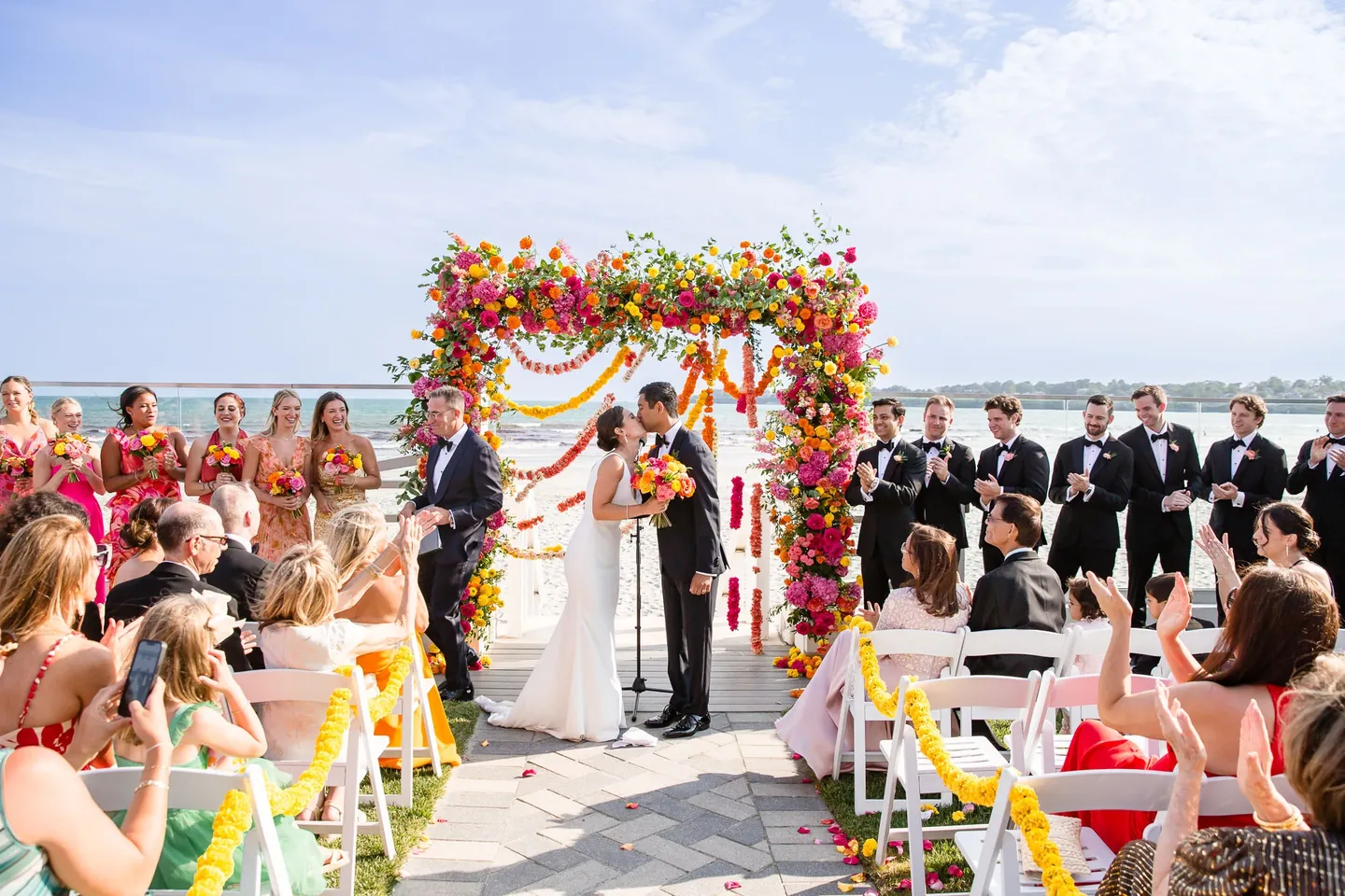 Wide view of the beachside ceremony, guests seated before a square arch heavy with bright flowers