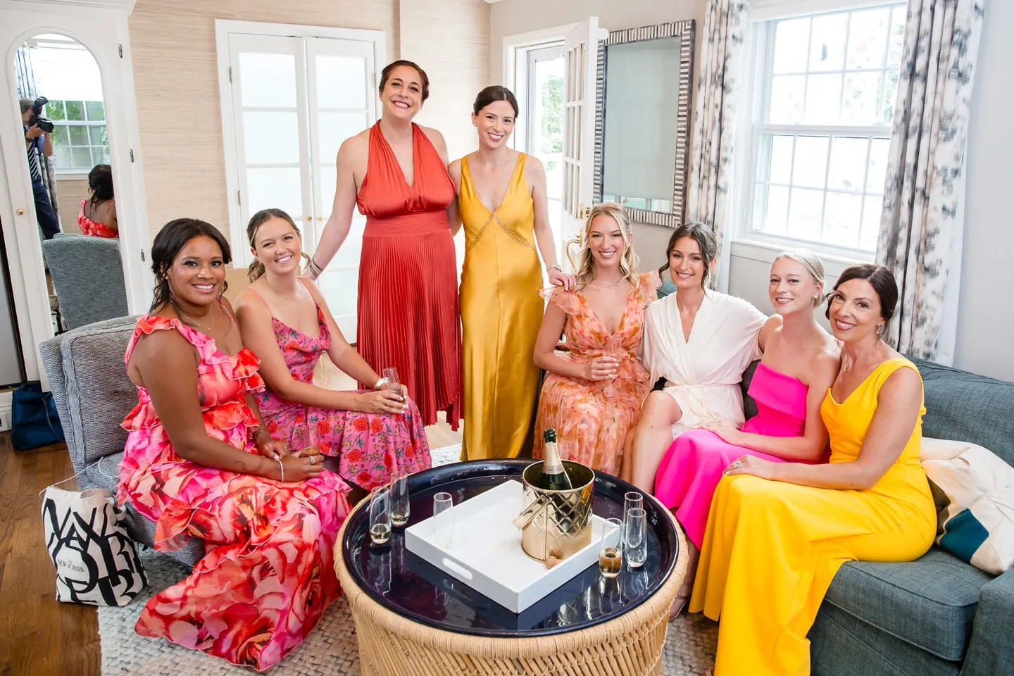 Bridesmaids in orange, fuchsia and gold gowns relax around the bride in a bright suite