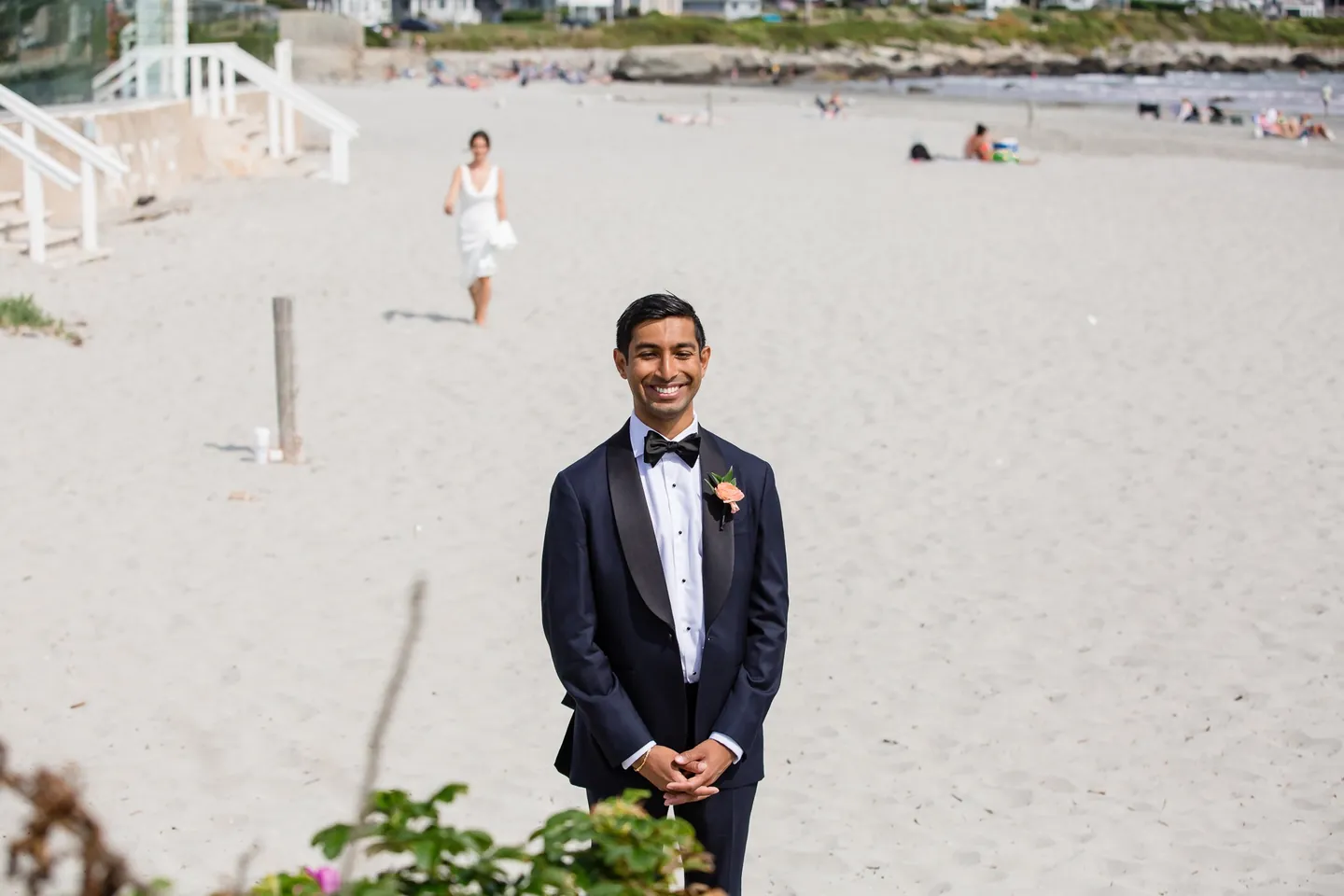 The groom waits on the beach with hands folded as the bride approaches across the sand behind him