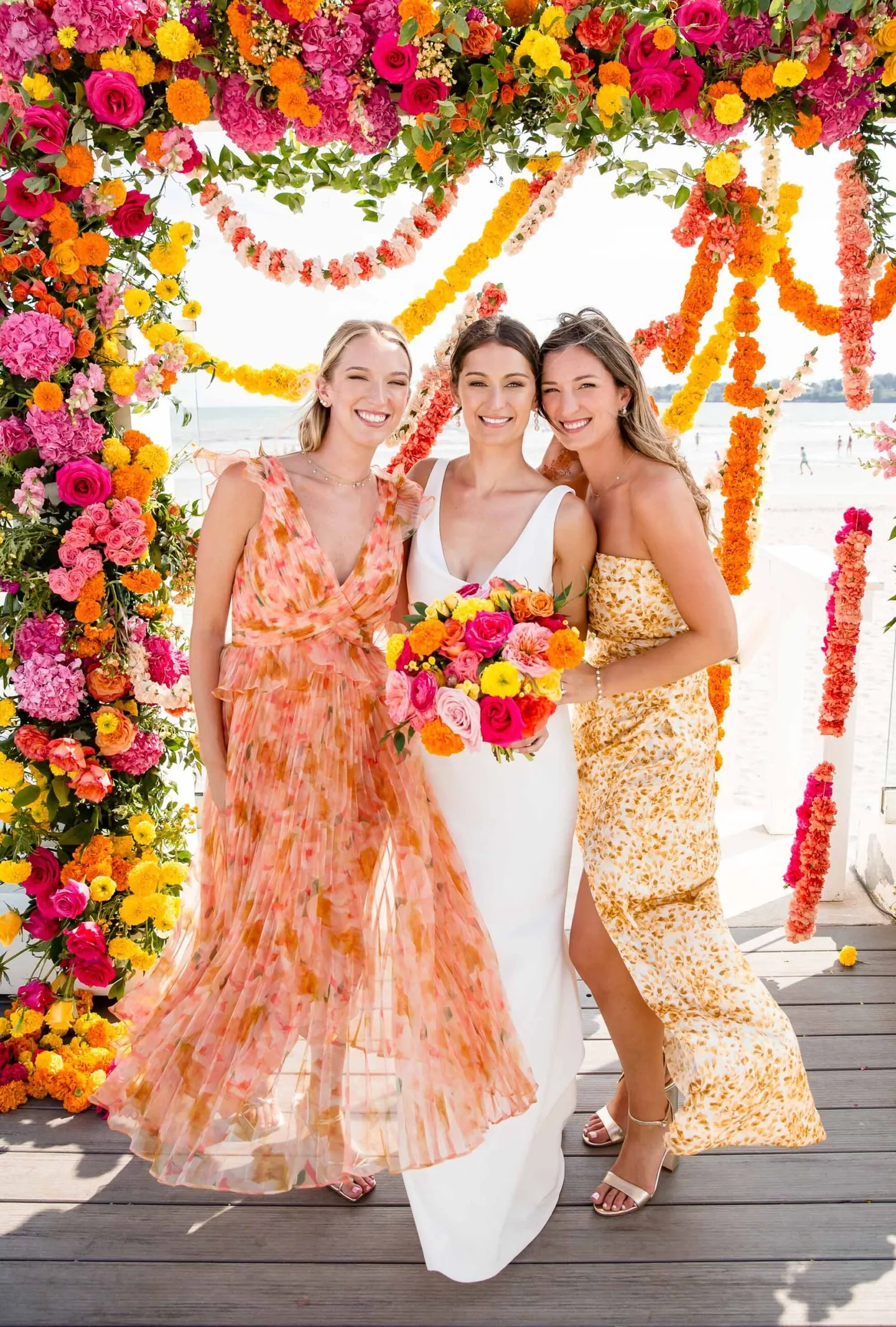 The bride with bridesmaids in peach and orange prints beneath the flower arch