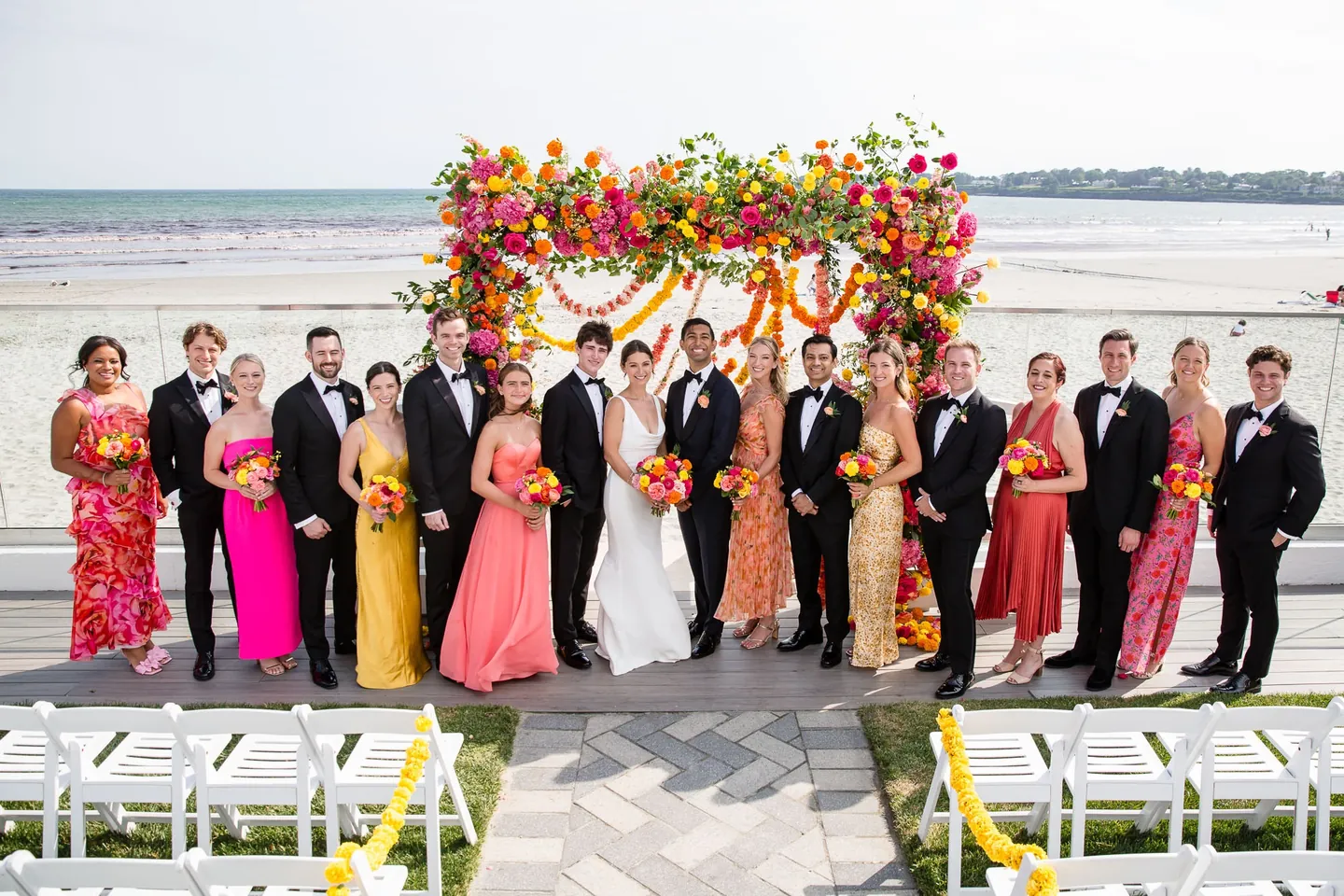 The full wedding party lines up before the floral arch with the beach and sky behind