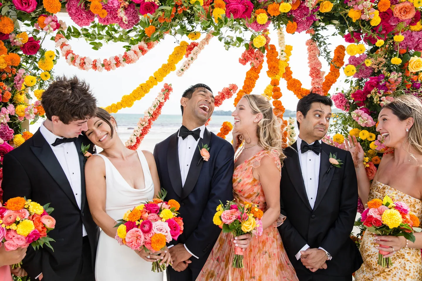 Groomsmen and bridesmaids burst out laughing around the couple under hanging garlands