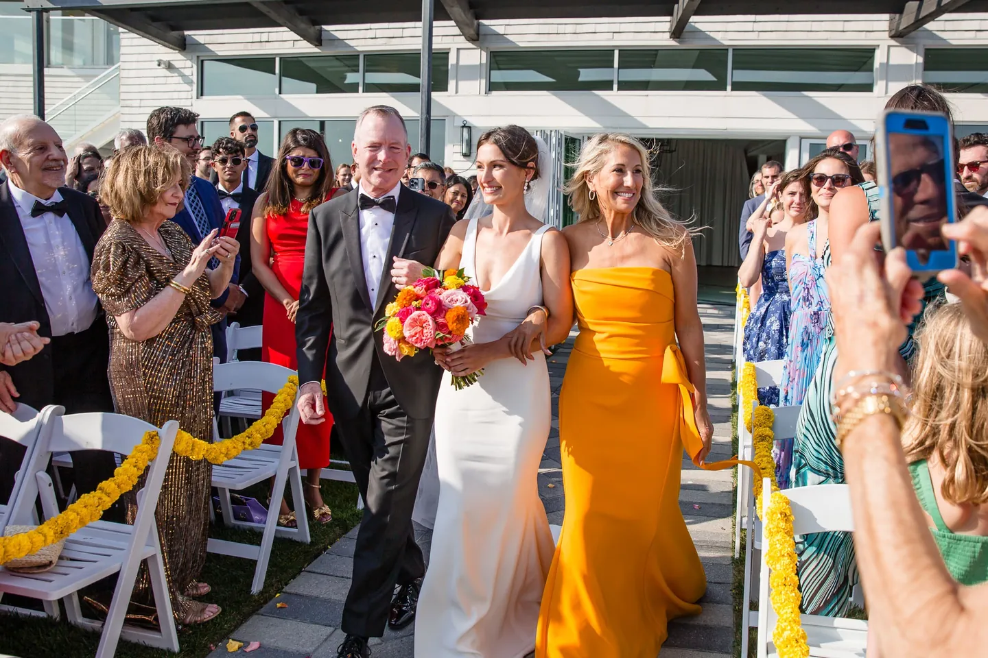 The bride walks the aisle between an older man in a tux and a woman in a saffron gown